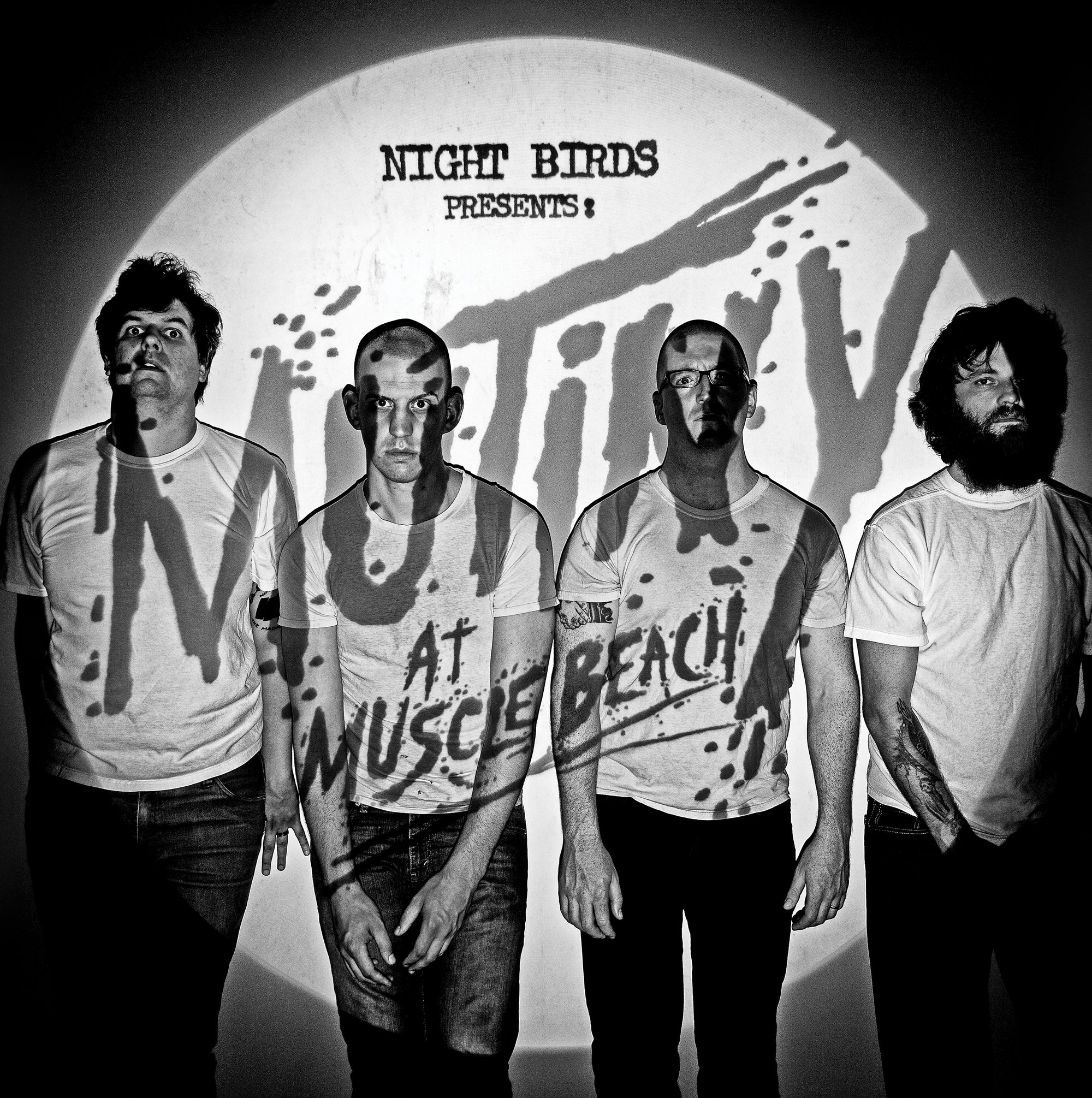 Four men stand before a wall with a Night Birds Presents Mutiny at Muscle Beach projection. The black and white shot, featuring each in plain white tees, reflects the raw energy of Night Birds’ Black Vinyl LP release.