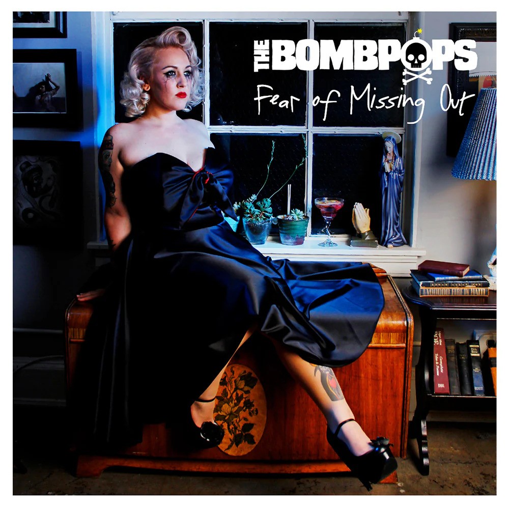 A blonde woman in a strapless black dress sits on a wooden chest in a vintage room. On the windowsill are a record, books, and drinks. Text: The Bombpops - Fear Of Missing Out (Brand: The Bombpops).