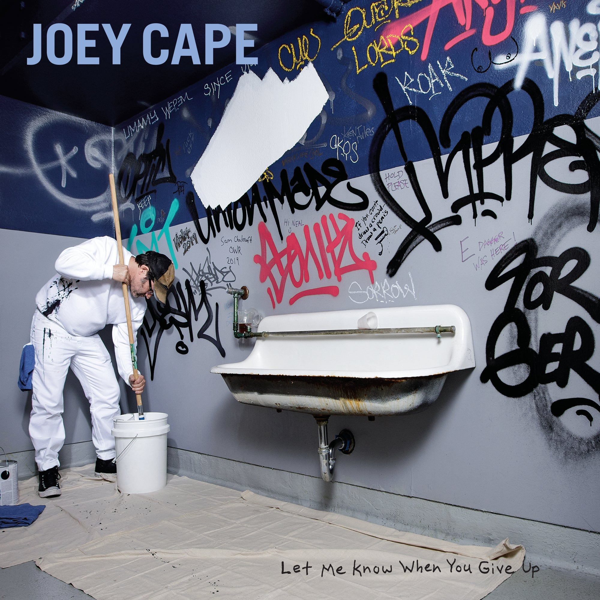 A man in white overalls paints graffiti off a blue wall above a sink. Joey Cape and Let Me Know When You Give Up are on the wall—artwork from the Joey Cape Black Vinyl LP release.